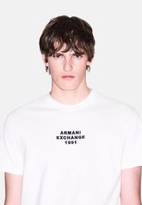 Biely bavlnený tričko s čiernym vyšívaným textom "ARMANI EXCHANGE 1991" na hrudi, krátke rukávy a okrúhly výstrih.