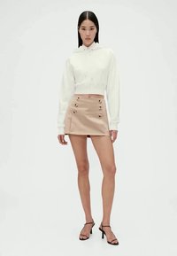 Cropped white hoodie with drawstrings, paired with a beige mini skirt featuring front buttons, and black high-heeled sandals.