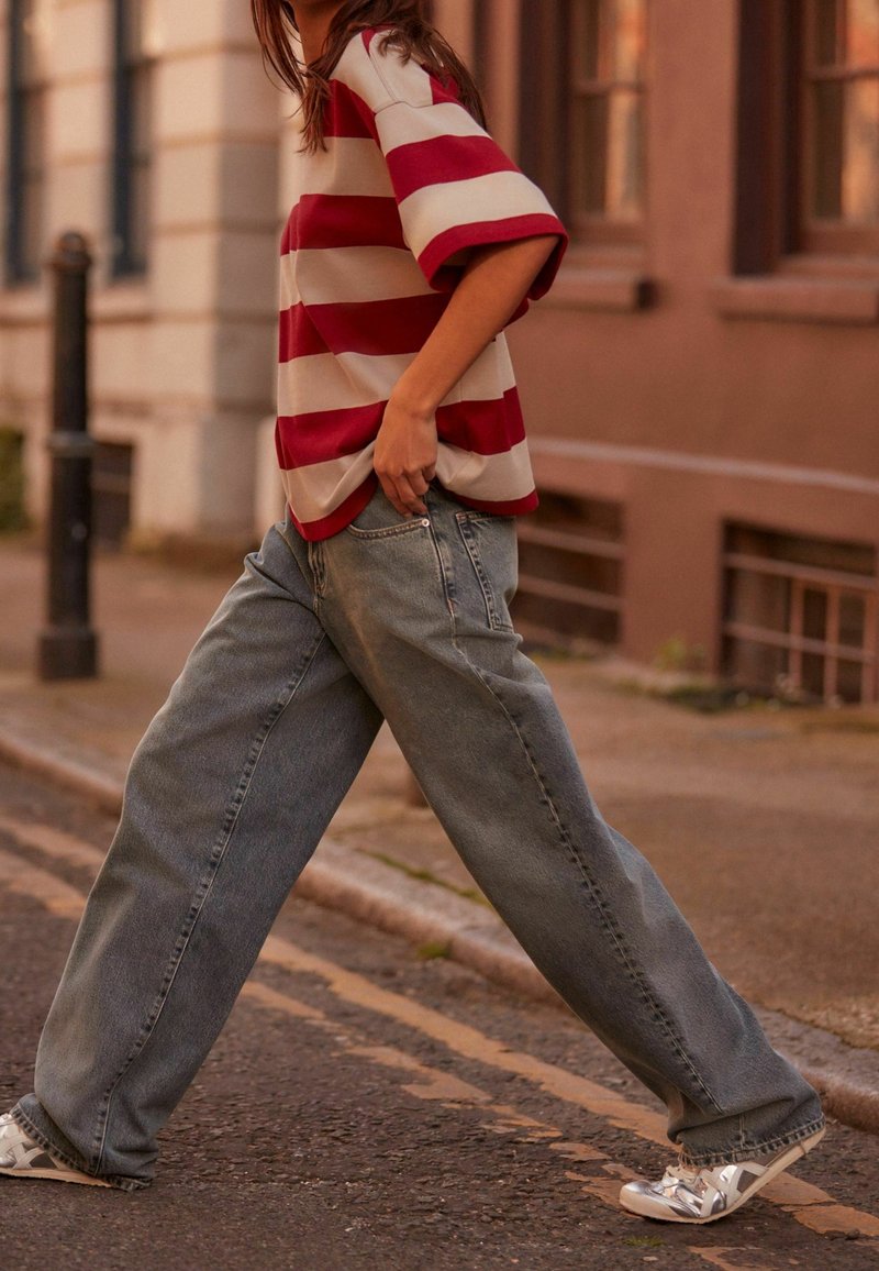 Chemise à rayures rouges et beiges à manches courtes, jeans en denim gris décontractés, baskets argentées avec des accents à motifs, marchant dans la rue.