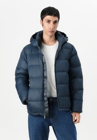 Navy puffer jacket with a hood, featuring a zipped front, quilted design, and logo accents. Paired with a white t-shirt and denim jeans.