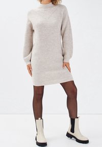 Femme portant une robe pull côtelée beige, des collants noirs transparents et des bottines Chelsea crème avec des élastiques noirs sur les côtés.