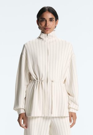 Nő krémszínű, vékony csíkos cipzáras dzsekiben, húzózsinóros derékkal és hozzáillő nadrágban, egyszínű háttér előtt áll.