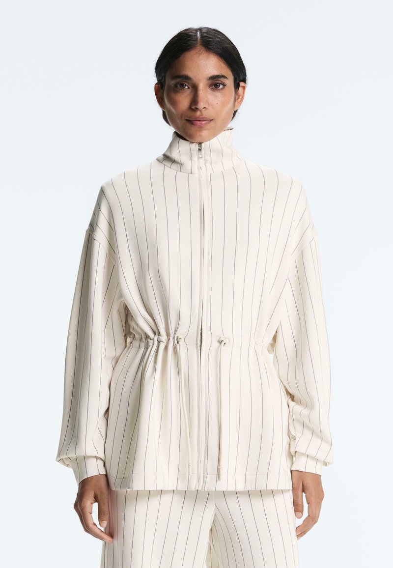 Ženska v kremno obarvani jakni s tankimi črtami na zadrgo in z vrvico v pasu ter ujemajočih se hlačah, stoji pred enobarvno ozadje.