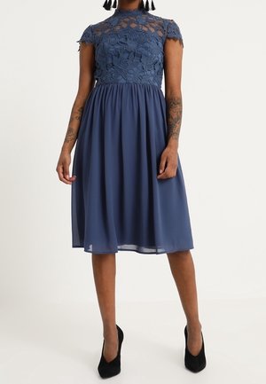 Woman wearing a knee-length navy blue dress with a floral lace bodice and short sleeves, paired with black pointed heels.