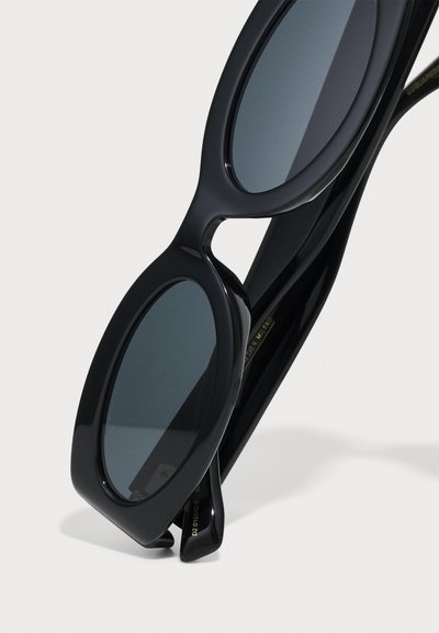Czarne owalne okulary przeciwsłoneczne z ciemnymi soczewkami, posiadające elegancką, błyszczącą plastikową ramkę i zwężające się zauszniki, umieszczone na sobie.
