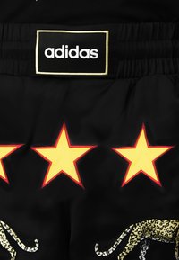 Melni sporta šorti ar baltu Adidas logo, izšūti dzelteni zvaigžņu motīvi ar sarkanu apmalīti un leopardu raksta aksentu.