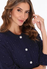 Cardigan lavorato a maglia blu navy con un motivo a onda testurizzato, arricchito da perline e bottoni simili a perle, con polsini arrotolati e scollatura rotonda.