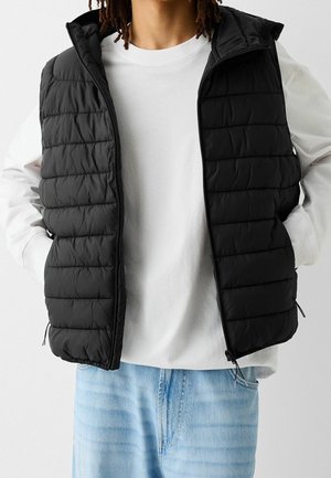 Person iført en sort quiltet ærmeløs vest over en hvid langærmet skjorte med lyseblå jeans, hænderne stukket i vestens lommer.