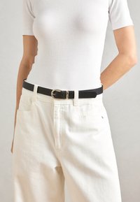 Ceinture en cuir noir avec une boucle en or, portée sur un pantalon blanc taille haute et une chemise blanche ajustée. Texture lisse, design minimaliste.
