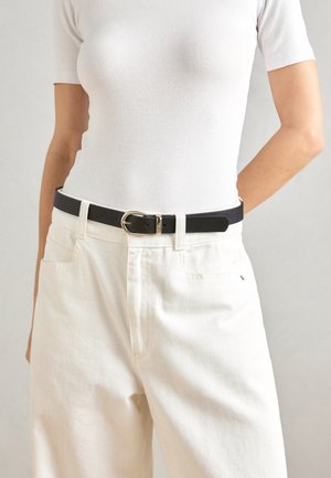 Cintura in pelle nera con fibbia dorata, indossata sopra pantaloni bianchi a vita alta e una camicia bianca aderente. Texture liscia, design minimalista.