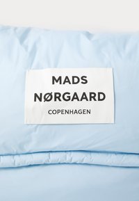 Gaiši zils audums ar gludu tekstūru, uz kura ir balta birka ar uzrakstu "MADS NØRGAARD KOPENHĀGEN" treknajiem melnajiem burtiem.