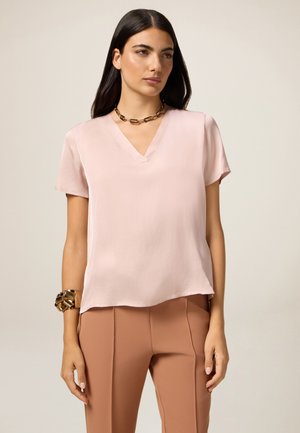Pink short-sleeve V-neck blouse in smooth fabric, featuring a relaxed fit and curved hem, paired with high-waisted beige trousers.