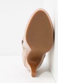 Chaussure à talon haut beige avec un extérieur en suède lisse, un bout pointu et une semelle en caoutchouc texturée avec des rainures horizontales.