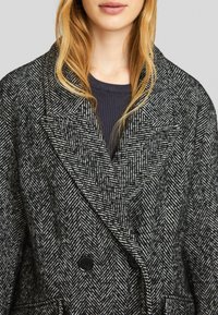Mantel im Fischgrätmuster in Schwarz und Weiß, mit einem doppelreihigen Design, großen schwarzen Knöpfen und breiten Revers.