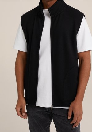 Man draagt een zwart mouwloos vest met ritssluiting over een wit T-shirt met korte mouwen en donkere spijkerbroek, staat met ontspannen armen.