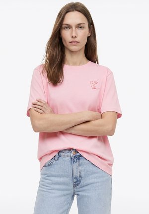 Woman with long brown hair wearing a pink t-shirt with a small red cat and heart design, standing with arms crossed, light blue jeans.