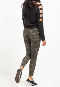 Czarny sweter z wycięciami na ramionach, w połączeniu z zielonymi spodniami typu skinny w kamuflażu i czarnymi trampkami z białymi podeszwami.