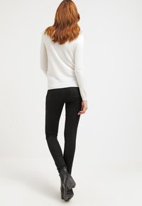 Vrouw met rood haar, gekleed in een witte longsleeve, zwarte skinny jeans en zwarte laarzen met hakken, staand met de rug naar voren gericht.