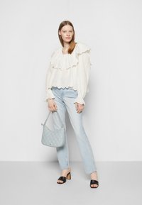 Blusa branca com pregas e detalhes em buracos, jeans azul claro, saltos abertos pretos e uma mala acolchoada azul claro. Modelo em uma pose de pé.