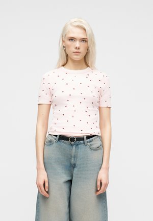 Blond kvinde iført en lyserød kortærmet skjorte med små røde hjerter, stoppet ned i højtaljede lyseblå vide bukser med sort bælte.