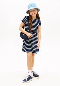 Κορίτσι που χαμογελά φορώντας μπλε και λευκό ριγέ φόρεμα, μπλε καπέλο κουβά, μπλε τσάντα χιαστί, λευκές κάλτσες και μπλε αθλητικά σε λευκό φόντο.