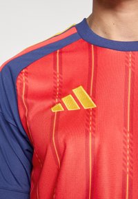 Röd sporttröja med marinblå axeldetaljer, prydd med gula ränder och en broderad gul Adidas-logotyp på bröstet.