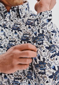 Biała koszula z bogatym niebieskim i granatowym wzorem kwiatowym, z zapięciem na guziki przy mankietach i klasycznym kołnierzykiem, prezentująca gładką fakturę materiału.