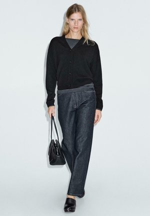 Cardigan nero con scollatura a V e bottoni, abbinato a jeans a gamba larga in denim scuro. Il modello porta una borsa nera e indossa zoccoli. Maglietta a righe visibile.