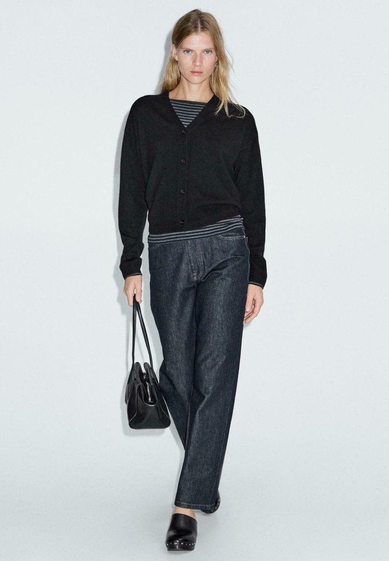 Svart cardigan med V-hals och knappar, tillsammans med mörk denim vidbyxor. Modellen bär en svart väska och har på sig träskor. Randig topp är synlig.