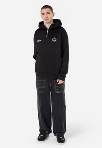 Felpa nera con cappuccio e collo con zip, decorata con ricami blu e dotata di tasca frontale. Abbinata a jeans neri larghi con cuciture dorate.