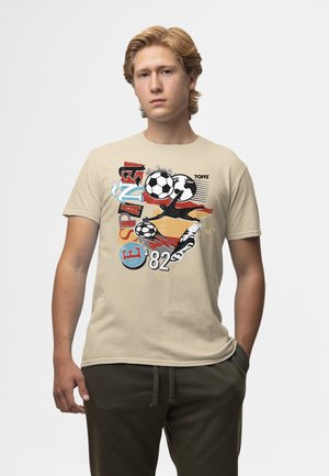 Ung mand iført beige T-shirt med grafik af fodboldbolde, målmandssilhuet, Spaniens flagfarver og teksten "ESPAN 82" og "TOFFS."