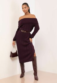 Off-shoulder kabelstrikket kjole i dyb burgunder med en tætsiddende silhuet, kombineret med brun bælte og knælange, tætsiddende brune støvler.