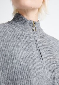 Gros plan d'une personne portant un pull tricoté gris avec un col montant et un tirette de fermeture éclair circulaire dorée.