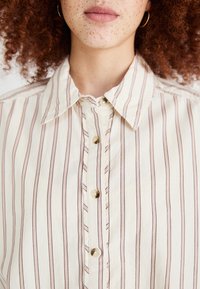 Camicia con bottoni realizzata in tessuto leggero, caratterizzata da strisce verticali crema e marroni, colletto a punta e bottoni rotondi.