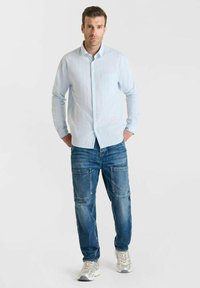 Man staat met handen in zijn zakken, gekleed in een lichtblauw overhemd met knopen, blauwe jeans met een opgestikte zak en witte sneakers op een witte achtergrond.