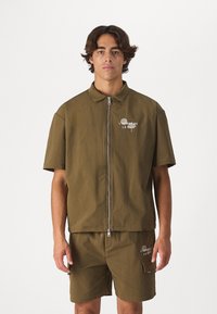 Olijfgroene kortekraagjacket met rits en een kraag, met witte tekst. Gecombineerd met bijpassende shorts, ook met tekstdetails.