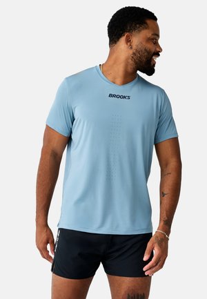 Mand iført en lyseblå Brooks sportst-shirt og sorte shorts, ser mod højre og smiler mod en ensfarvet baggrund.