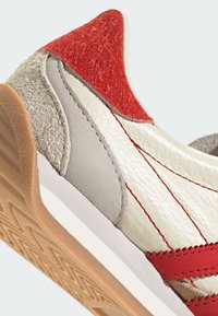 Retro sneakers featuring a beige leather body, red and gray accents, textured suede, and a rubber sole with a geometric pattern.