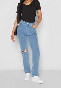 Person som bär en svart V-ringad T-shirt och höga ljusblå jeans med ett rivet knä, håller en svart väska. Avslappnad urban stil.