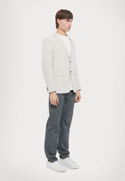 Helle graue Blazer mit zwei Knöpfen und seitlichen Taschen, kombiniert mit dunklen Jeans und weißen Sneakern. Einfaches Design, strukturiertes Material.