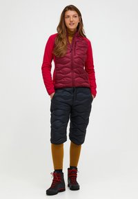 Vrouw staat met handen in de zakken, draagt een rode jas, zwarte gewatteerde shorts, mosterdkleurige leggings en zwarte wandelschoenen met rode veters.
