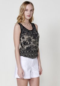 Débardeur avec un motif floral noir sur fond beige, encolure arrondie et bord noir. Associé à un short blanc. Texture de tissu lisse.