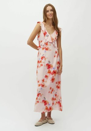 Młoda kobieta ubrana w jasnoróżową, bezrękawnikową sukienkę z czerwonymi i fioletowymi kwiatowymi wzorami oraz falbankami, w połączeniu z beżowymi, plecionymi balerinami.