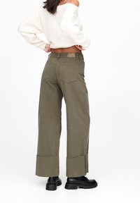 Pantalon large vert armée en coton avec ourlet brut et deux poches arrière, associé à des bottes noires épaisses et un pull blanc court.