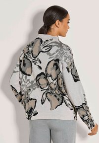 Frau mit dunklem Haar in einem Pferdeschwanz, die einen lässigen grauen Pullover mit großen abstrakten Blumenmustern und hellgrauen Hosen trägt, mit dem Rücken zur Kamera.