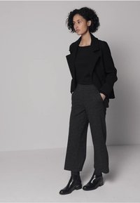 Musta villasekoitebleiseri, jossa on jämäkät olkapäät, yhdistetty hyvin istuviin tummanharmaisiin villahousuihin. Kiiltävät mustat nilkkurit viimeistelevät asun.