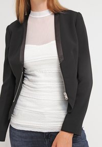 Giacca nera con zip e rifiniture in satin, indossata sopra una maglietta bianca strutturata con righe orizzontali e scollatura trasparente.