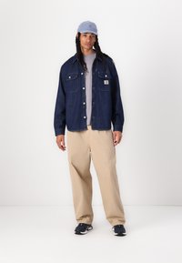 Denim jacket in dark blue with two chest pockets, gray t-shirt underneath, beige trousers, and light blue cap. Black sneakers complete the look.