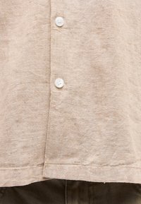 Κοντινό πλάνο μπροστινού μέρους μπεζ λίνεν πουκάμισου με δύο λευκά κουμπιά και ορατή ραφή πάνω από σκούρο παντελόνι από ύφασμα.