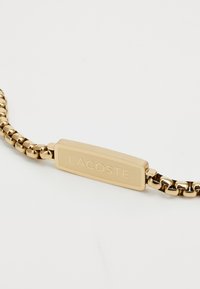 Bracelet en chaîne dorée avec une plaque rectangulaire lisse gravée "LACOSTE," reliée par des segments métalliques arrondis.